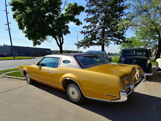 Delivered: gold metallic classic luxury coupe with a textured vinyl landau roof and opera windows, parked on the lot beside other classics