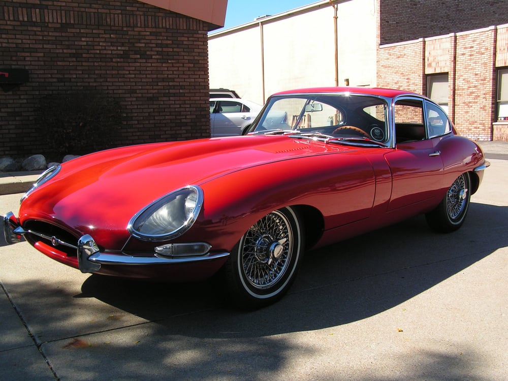 Delivered: bright red Jaguar E-Type fixed-head coupe with chrome wire wheels and whitewall tires in the sun